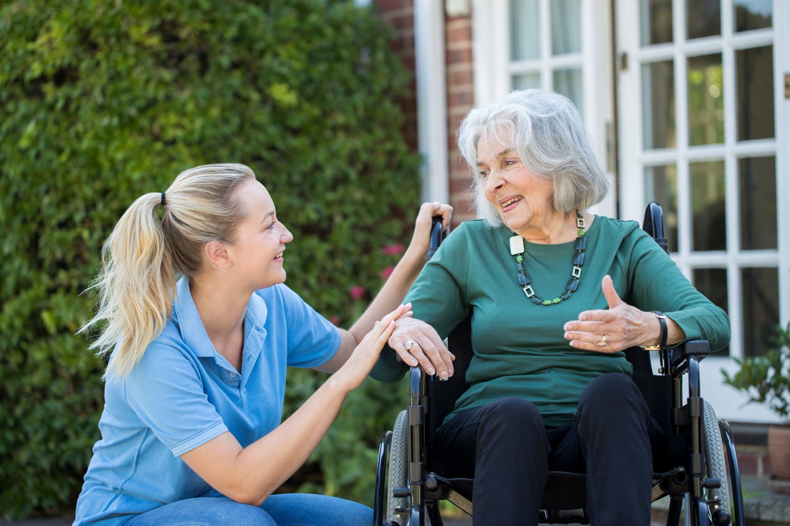 A caregiver kneeling beside a smiling client in her wheelchair outside her home